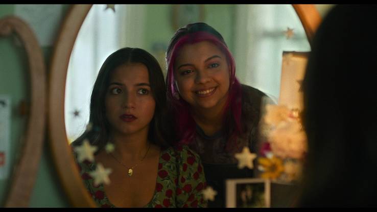 Aza and Daisy stare into the mirror in a bedroom. Daisy is smiling while Aza looks at her questioningly.