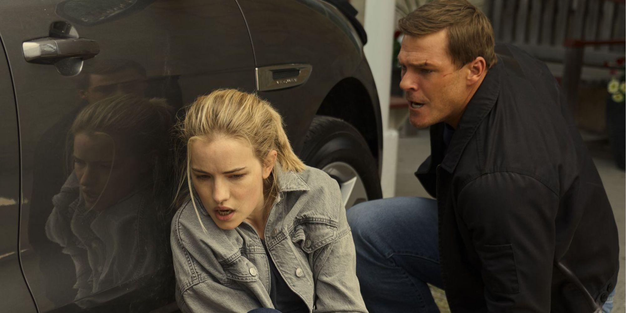 Willa Fitzgerald as Roscoe Conklin hiding behind a car with Alan Ritchson's Reacher.