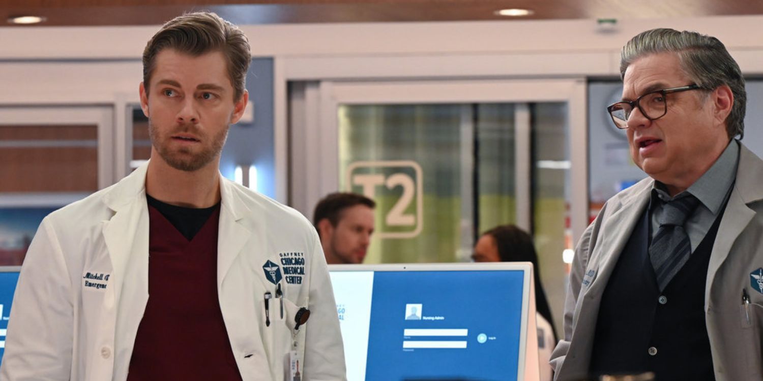 Chicago Med Ripley and Charles stand facing the camera as if staring at something in the distance. There is a computer screen behind them with a blue log in screen and two doctors behind it by Treatment Room 2, whose label is clearly visible behind the computer.