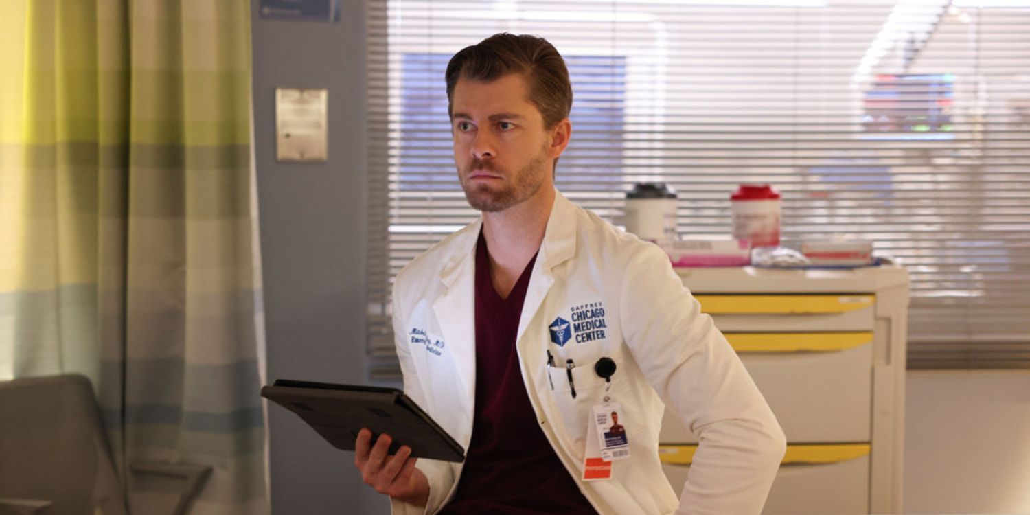 Chicago Med Ripley stands facing the camera, holding a tablet in his right hand and wearing a lab coat. There is a yellow plaid curtain to his left and behind him there is a counter with yellow stripes and jars on top and open blinds behind the counter, looking out on a yellow building with a window.