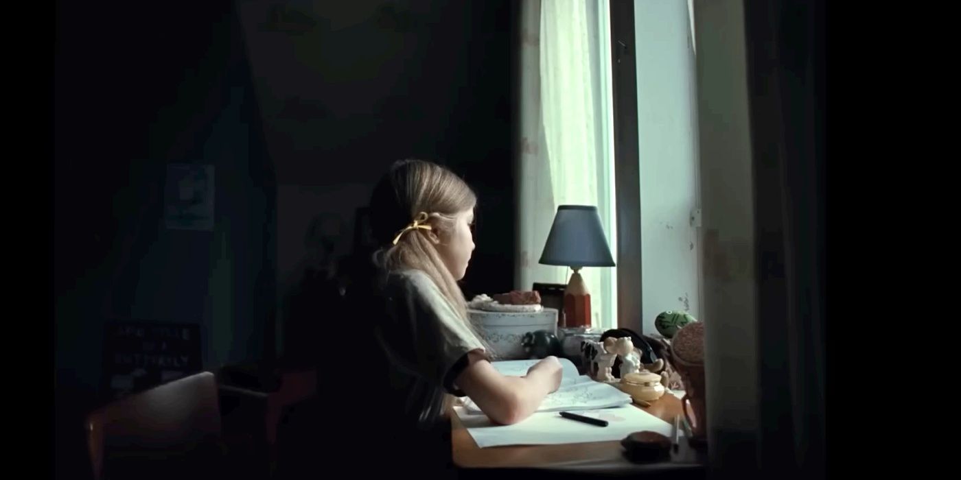 Uma menina olhando pela janela enquanto está sentada em uma mesa em Longlegs.