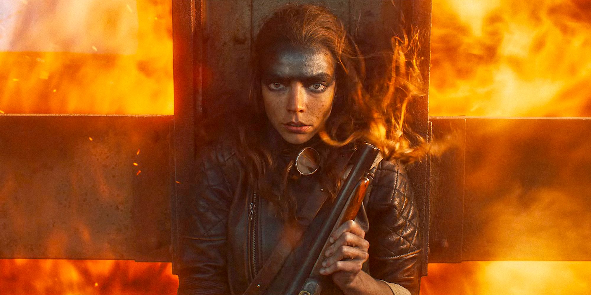 Furiosa, played by Anya Taylor-Joy, hiding behind a post from an explosion of flames. She is holding a gun, with her long hair blowing. Her face is painted with black oil.