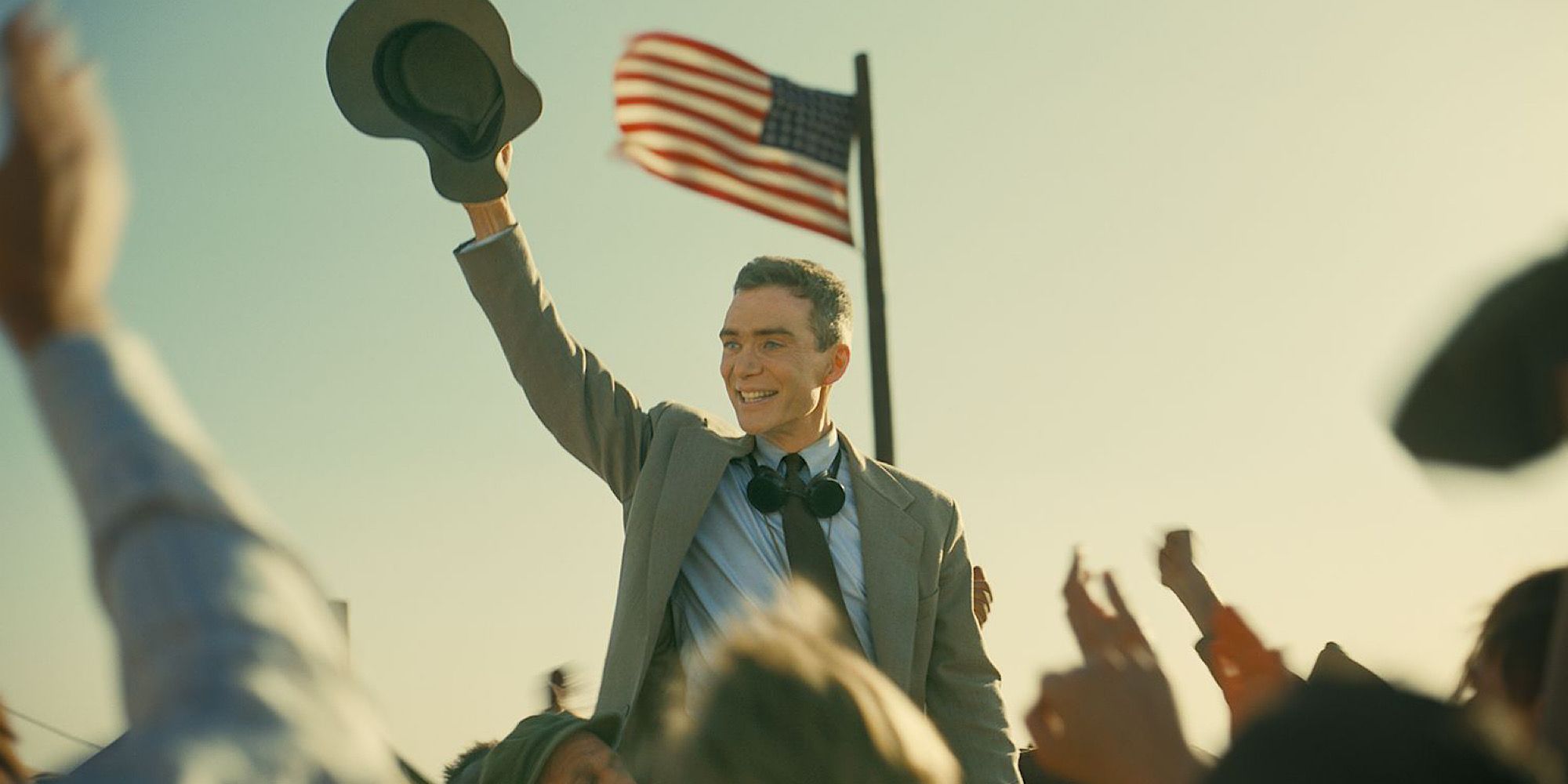 Cillian Murphy as Oppenheimer celebrating above a cheering crowd with the American flag in the background.