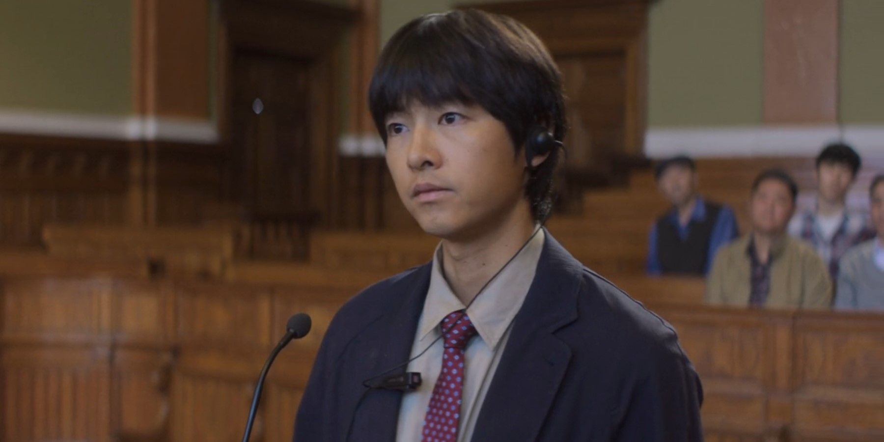 Loh Kiwan (Song Joong-ki) wears a translator ear piece and stands in front of a microphone during his trial to determine if he applies for Belgian refugee status in My Name is Loh Kiwan