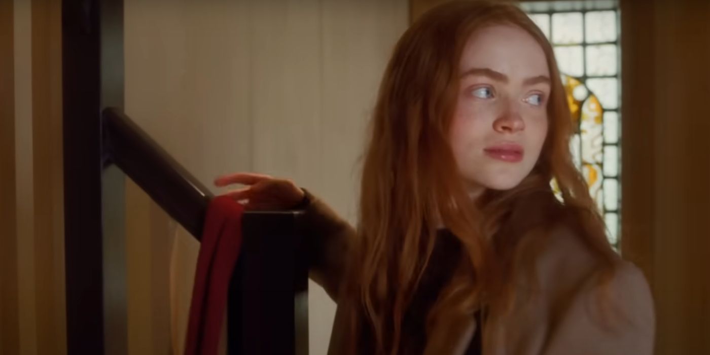 Sadie Sink in All Too Well with her hand on her red scarf draped over a stair bannister