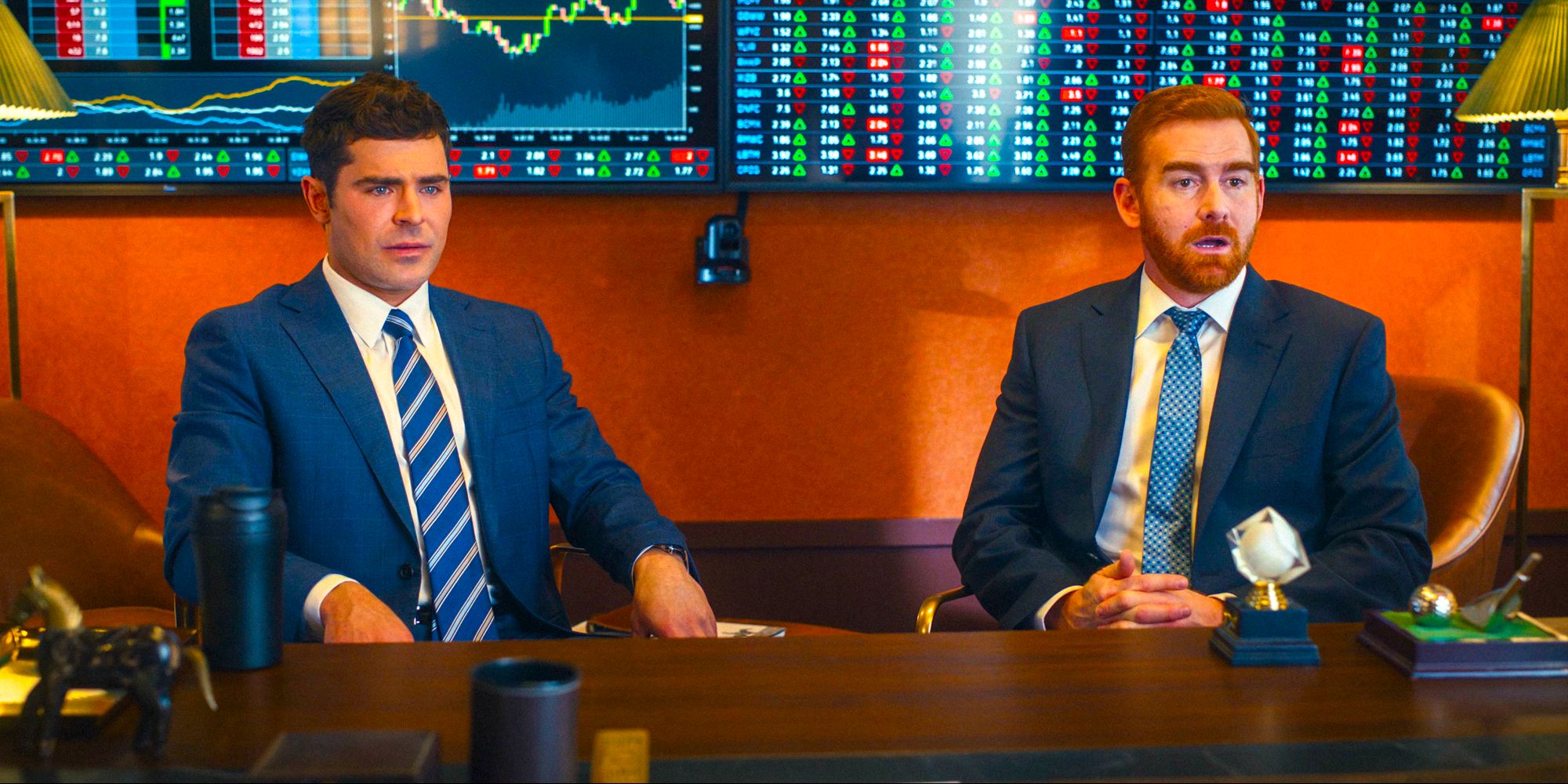 Dean and JT in suit sitting in front of charts in Ricky Stanicky
