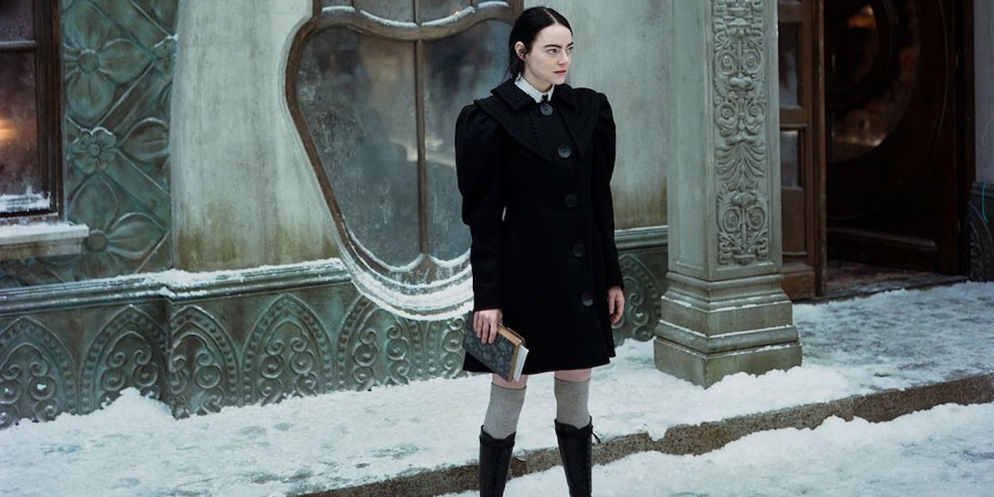 Bella Baxter (Emma Stone) stands on the snowy street in a black coat and knee high socks and boots in Poor Things.