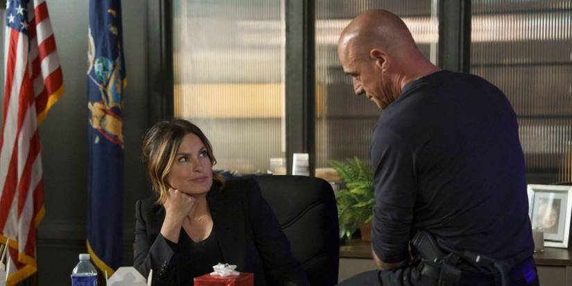 Benson sits in her desk chair, leaning her jaw on her right hand and looks up at Stabler, who is sitting on the edge of her desk to her right. There is a glass window behind Benson and an American flag to her right. There is a gift box on her desk.