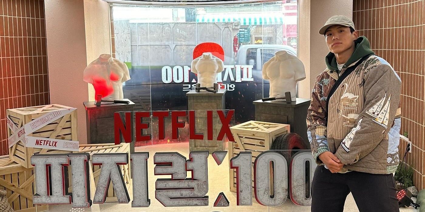 Amotti da 2ª temporada do Physical 100 posa com as mãos cruzadas em frente à placa do Netflix Physical 100 em coreano.