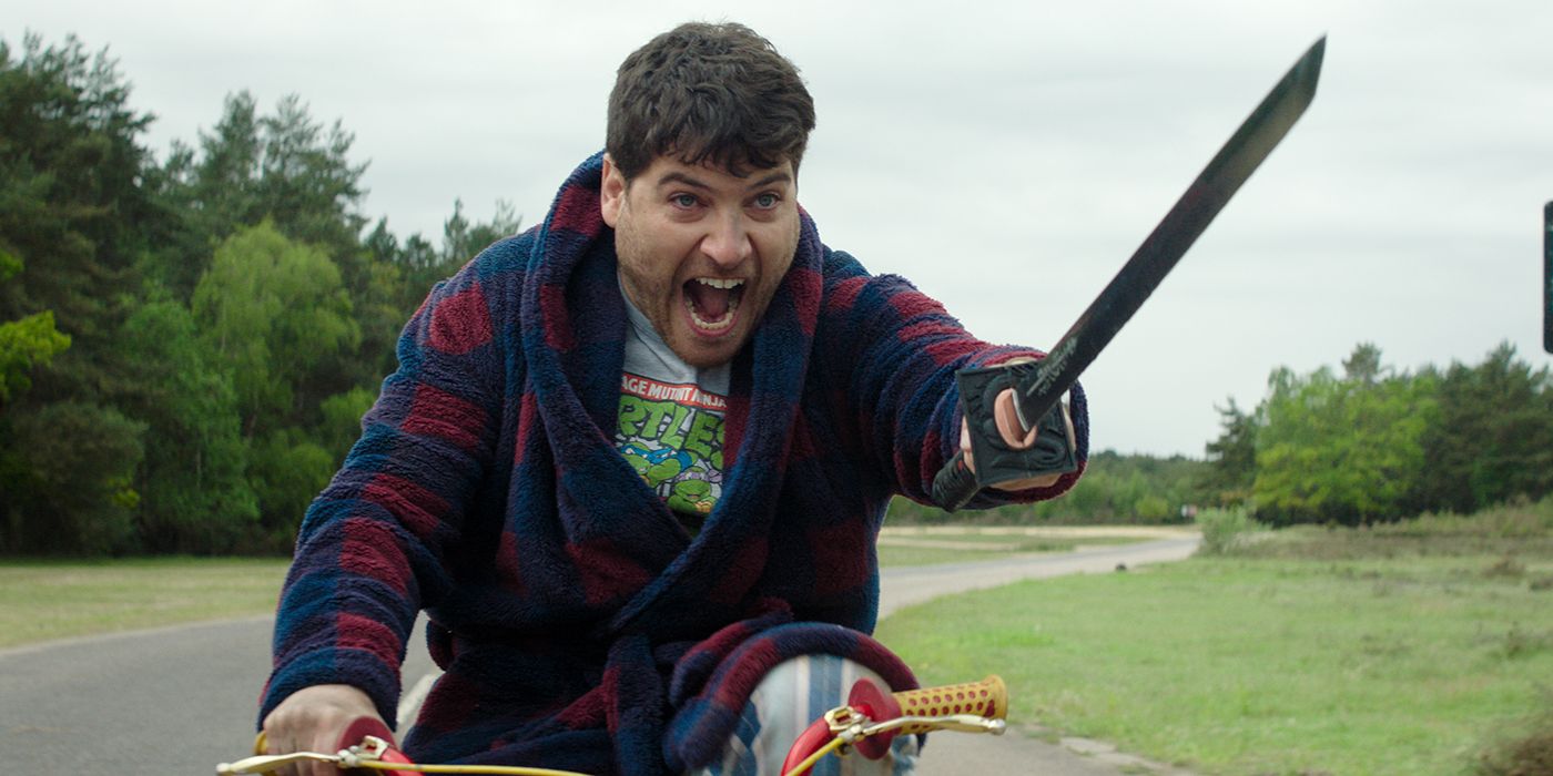 Adam Pally como Wade gritando enquanto andava de bicicleta e empunhava uma espada em Knuckles
