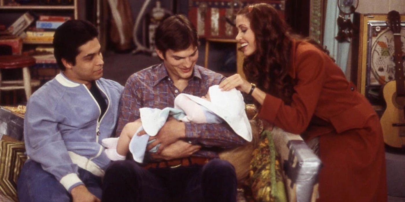 Kelso (Ashton Kutcher) sits next to Fez (Wilmer Valderrama) and Brooke (Shannon Elizabeth) while holding his baby daughter Betsy in That '70s Show