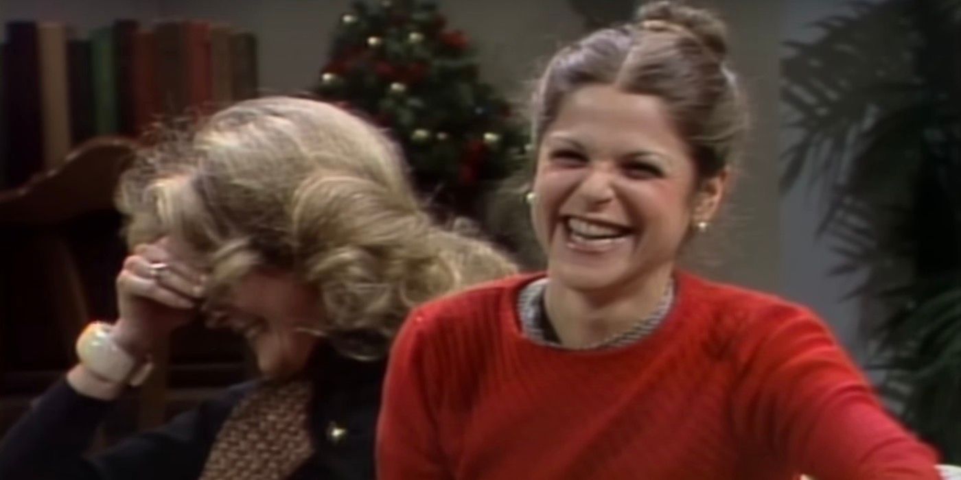 Candice Bergen and Gilda Radner are sitting next to one another on a couch and laughing.