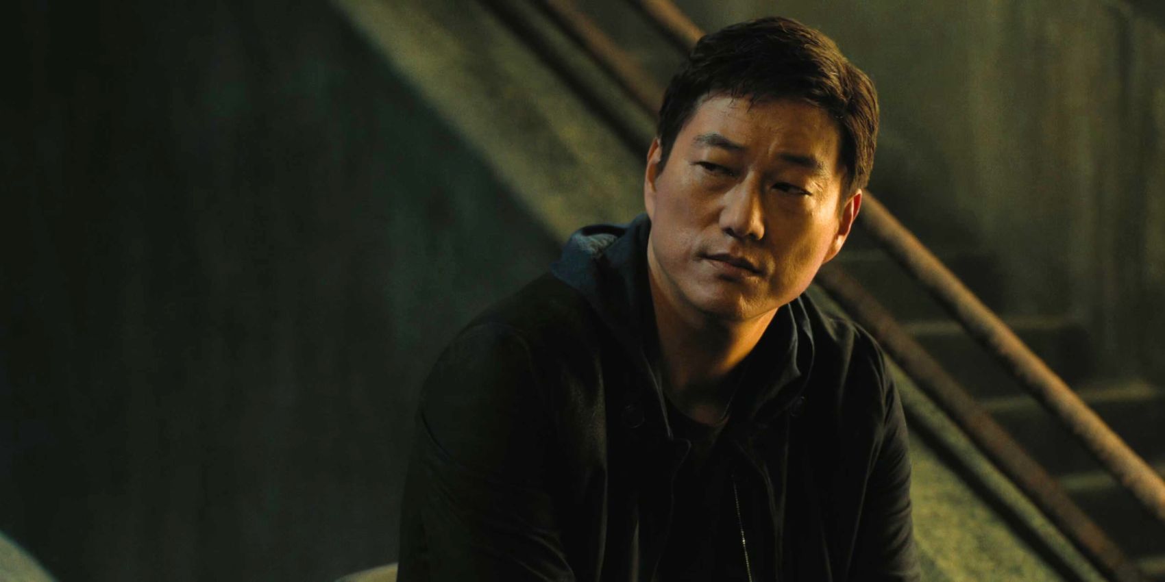 Han Lue seated in front of stairs with a somber expression in F9