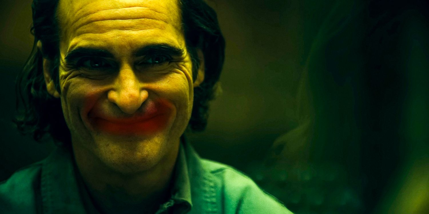 Arthur Fleck (Joaquin Phoenix) with a forced smile perfectly aligns with a red lipstick-drawn smile on a glass in Joker: Folie à Deux