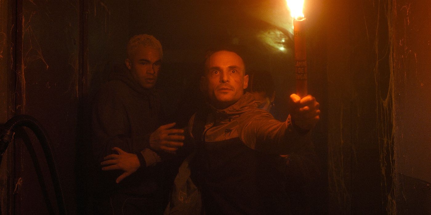 Mathys holding a flare in front of him and Kaleb in a dark hallway with spider webs in Infested