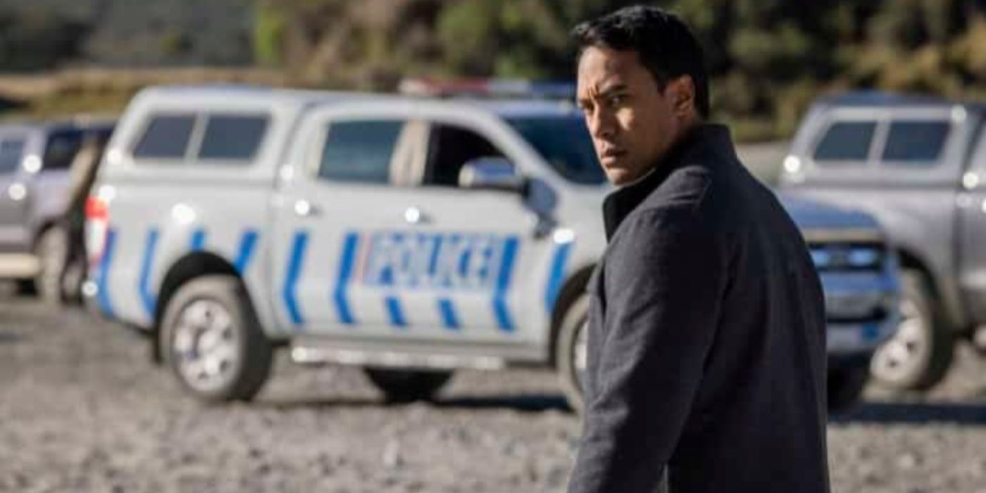 Dominic Ona-Ariki como Ariki Davis parece sério enquanto explora uma cena de crime do lado de fora, na frente de carros de polícia em One Lane Bridge
