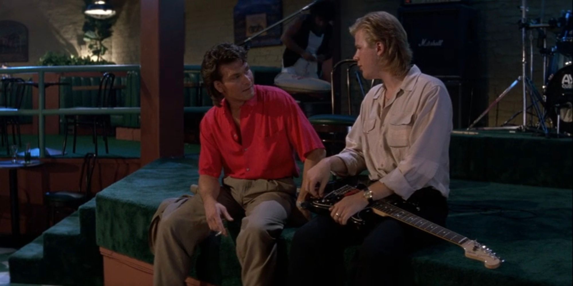 Dalton (Patrick Swayze) and Cody (Jeff Healey) sit at the edge of the stage and talk in Road House (1989).