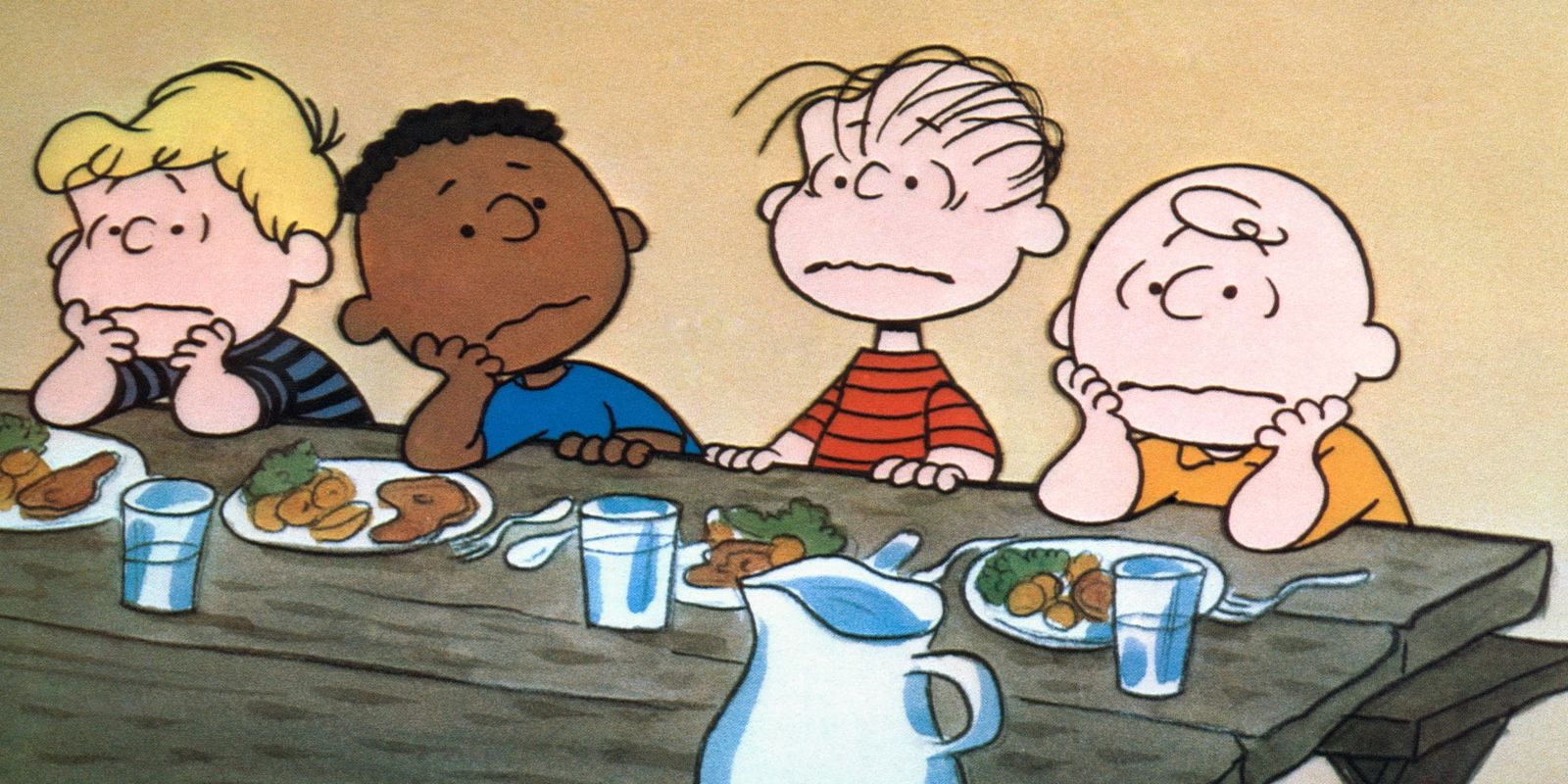 Peanuts kids sitting at a wooden table with food on plates in front of them.