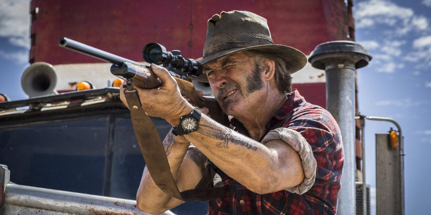 John Jarratt as Mick Taylor aiming his gun at something while standing on a truck in Wolf Creek 2 (2013) 