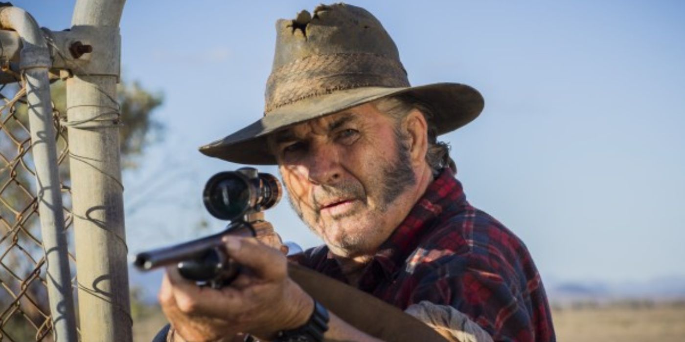 Mick Taylor apunta con su arma a alguien mientras está parado junto a una valla en Wolf Creek 2 (2013)