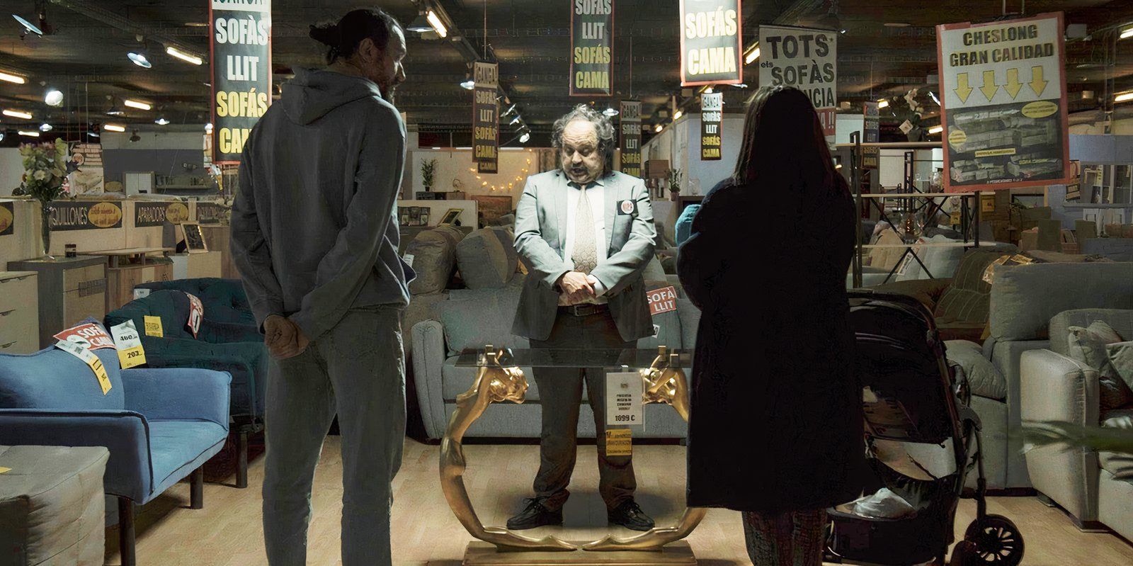 a couple standing in the middle of a furniture store with their backs to the camera, facing a salesman looking down at an ornate coffee table in The Coffee Table