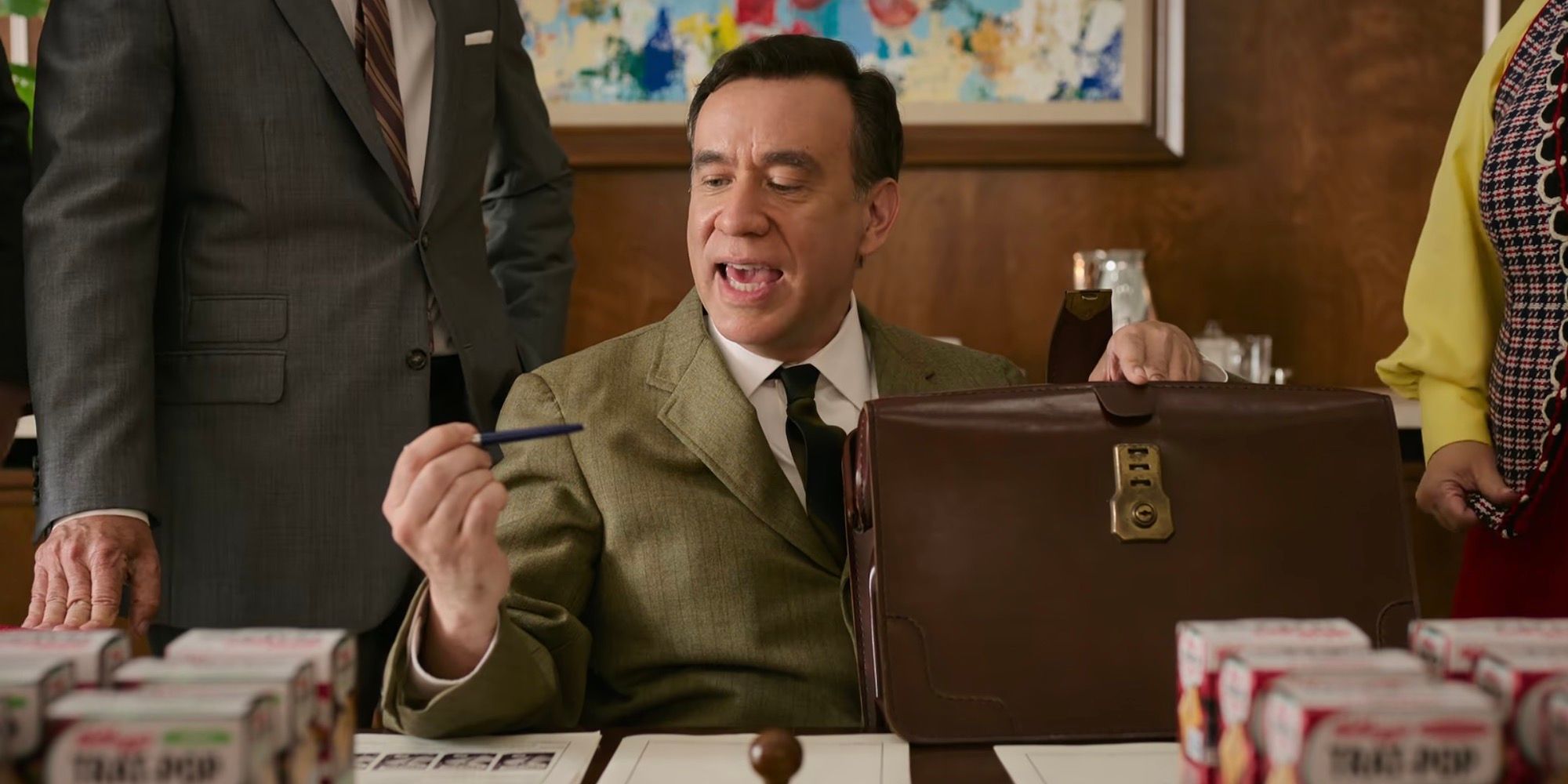 Fred Armisen is sitting down with a briefcase in one hand and a pen in the other. 