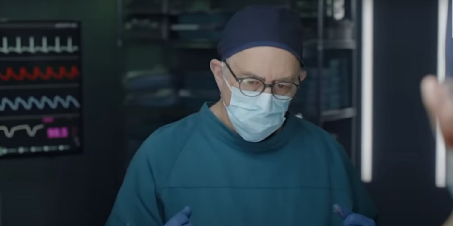 O Bom Doutor Glassman congela durante a cirurgia.  Ele está usando uniforme e há um monitor atrás dele registrando os sinais vitais do paciente.