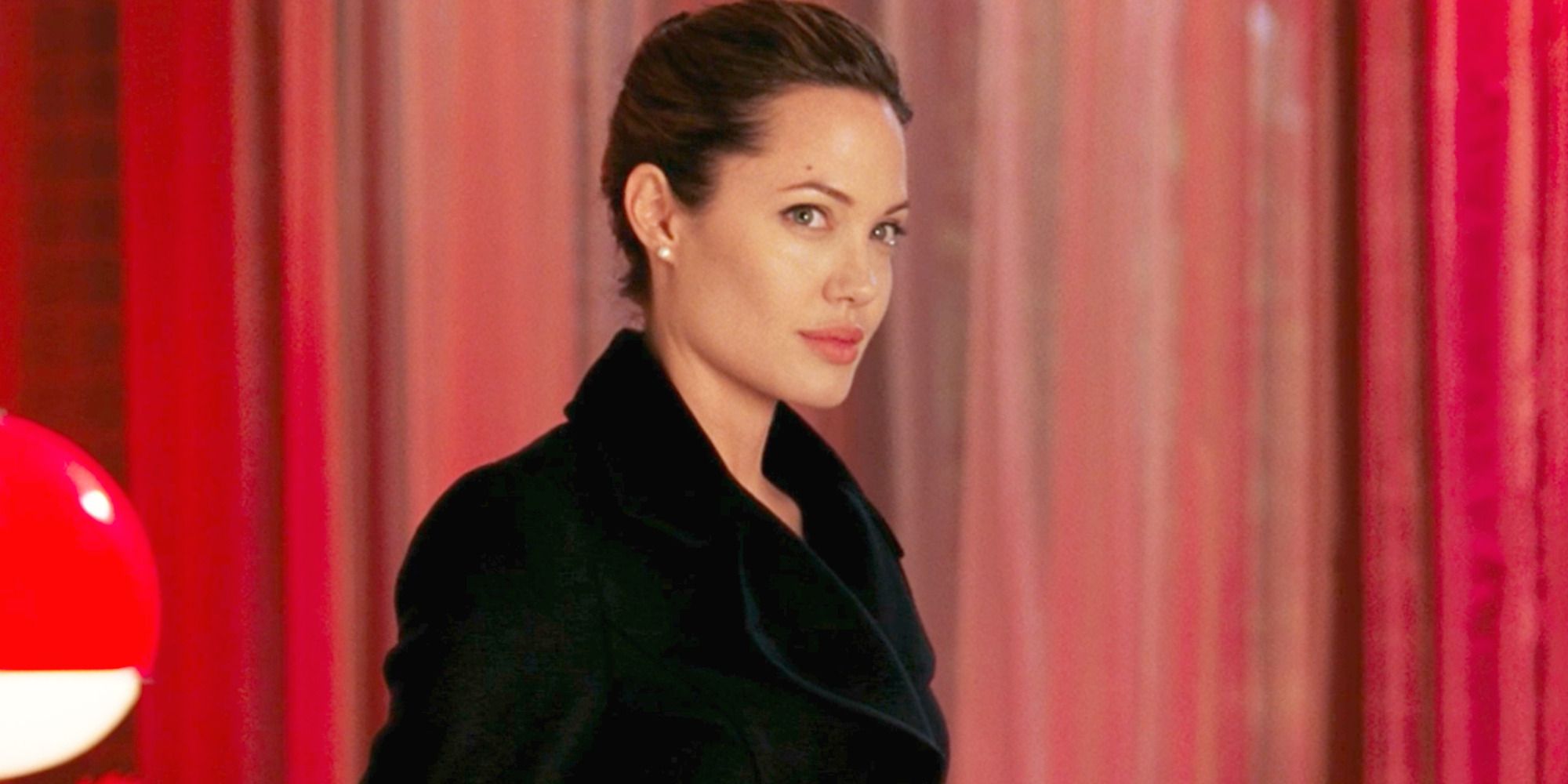 Jane (Angelina Jolie) causa problemas para o Sr. e a Sra. Smith enquanto usa um casaco preto em frente a uma cortina vermelha