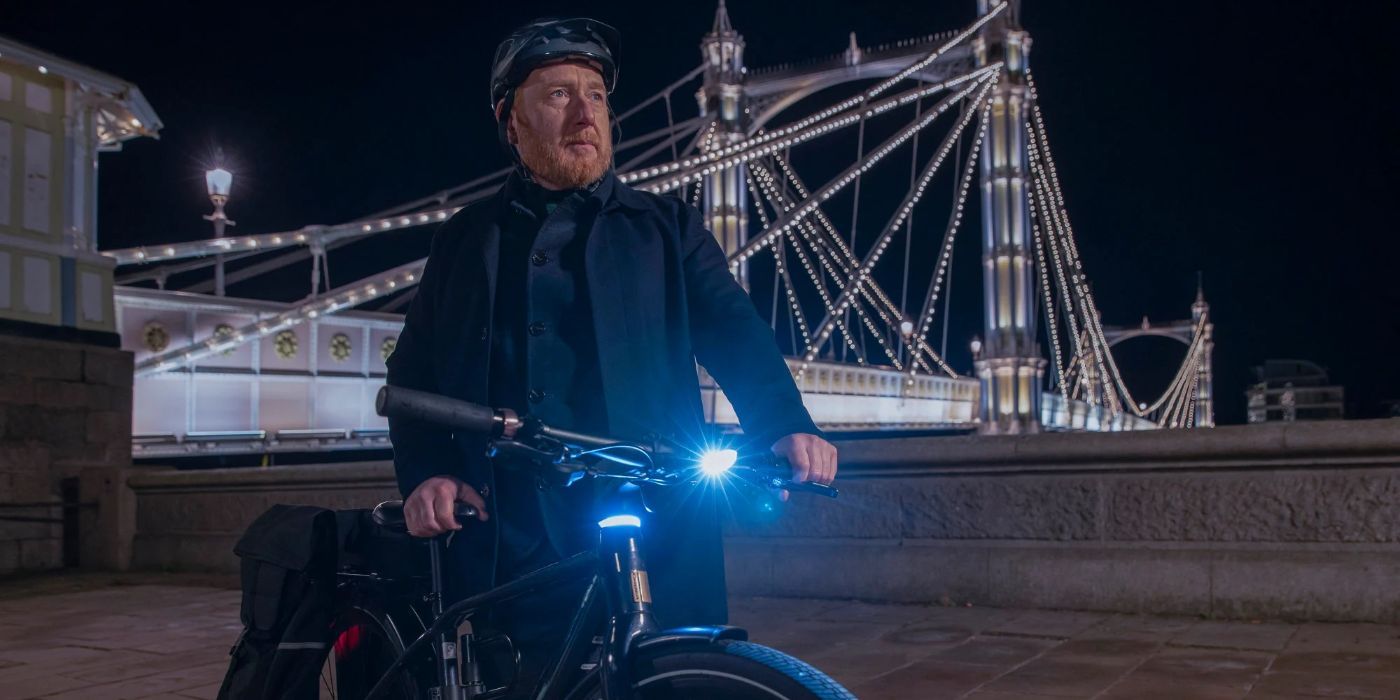 Max (Adrian Scarborough) parado ao lado de sua bicicleta perto de uma ponte à noite em The Chelsea Detective.