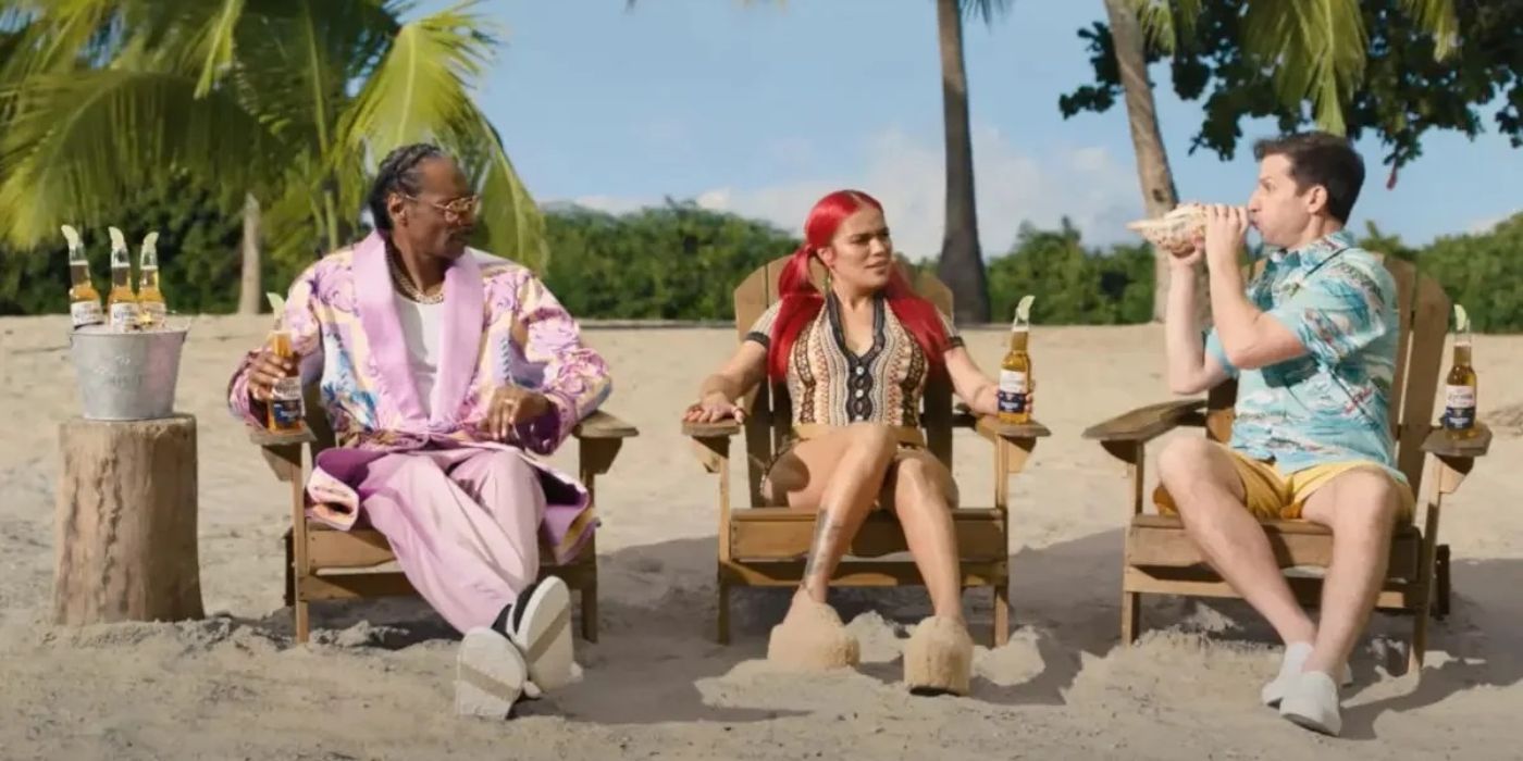 Snoop Dogg and Karol G looking at Andy Samberg blow a conch shell on the beach in a Corona commercial.