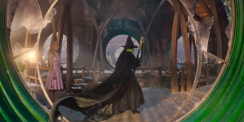 Elphaba (Cynthia Erivo) in her witch costume holding her magic broom, seen through a circular window with broken glass in Wicked