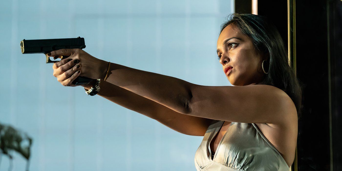 Shalini Peiris points a gun offscreen while wearing a silver dress