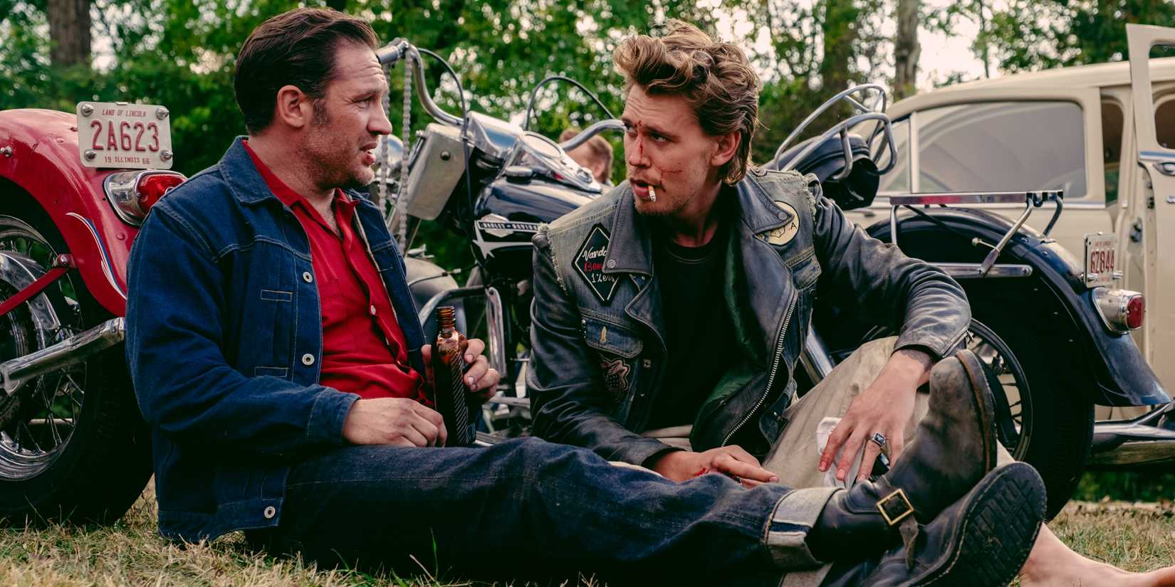 Johnny and Benny sitting on the ground talking in front of motorcycles in The Bikeriders