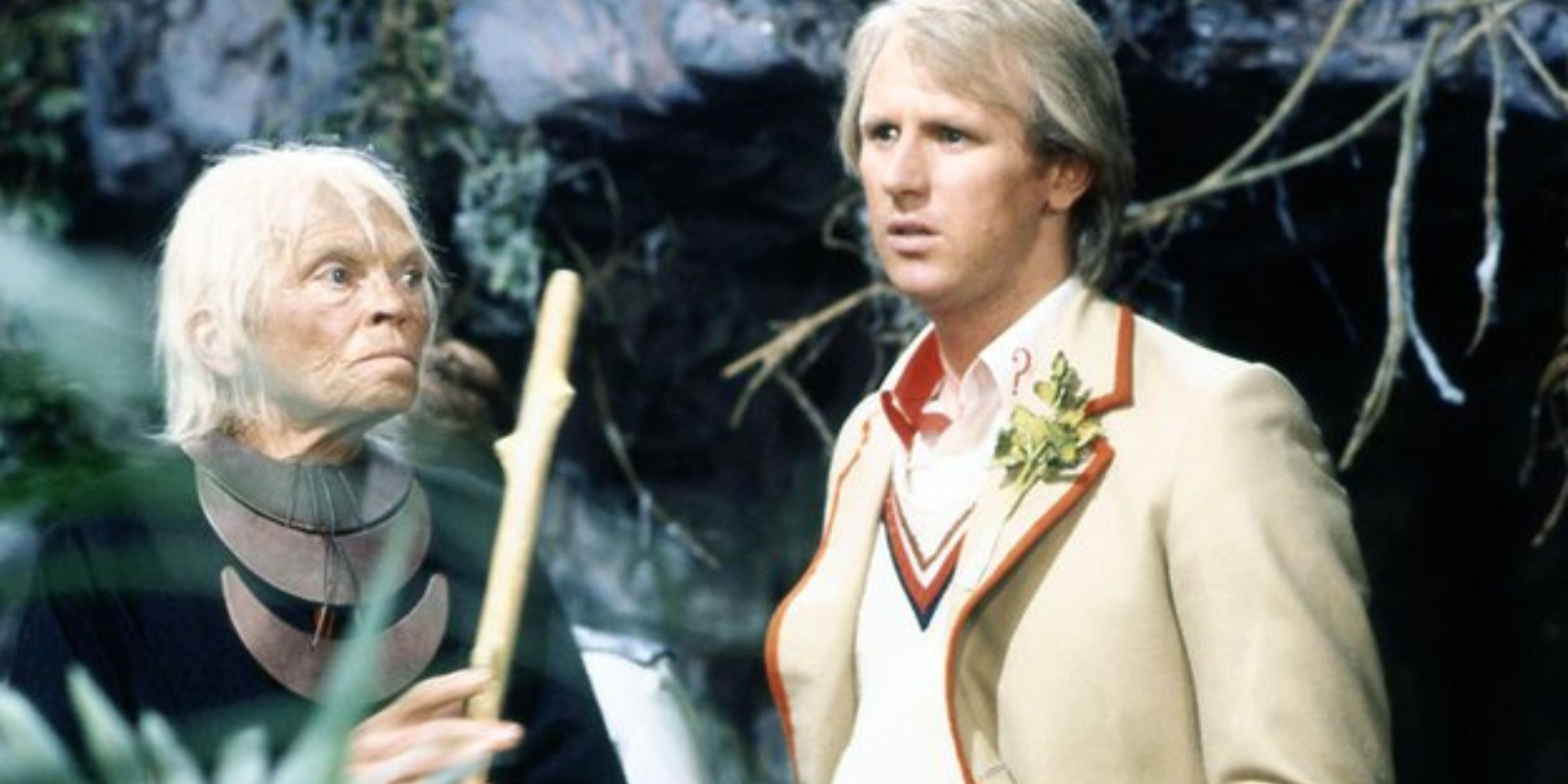 Peter Davison's Doctor standing next to an older woman holding a stick on Doctor Who
