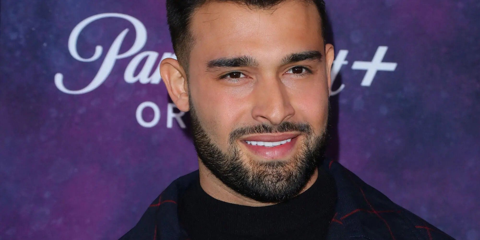 Sam Asghari sorrindo em um tapete vermelho com fundo roxo.