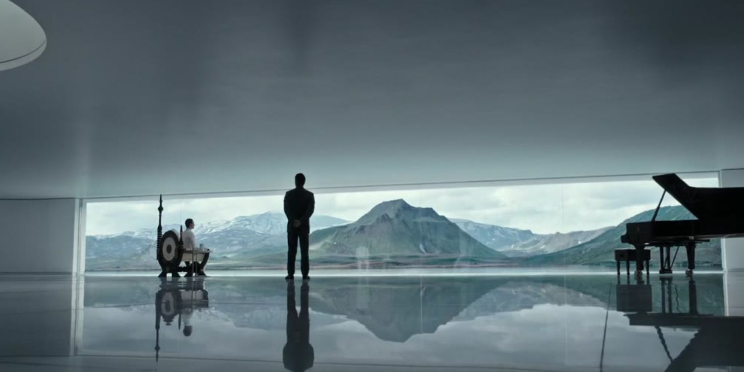 A still from the introductory sequence to Alien: Covenant. In a large room with a mirrored, spotless floor, Weyland is in a wheelchair overlooking mountainous scenery beneath a blind. David is standing next to him with his hands behind his back.