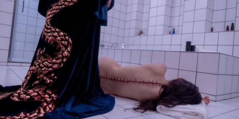 A still from The Substance. In a stark, white-tiled bathroom, a naked body (Elisabeth) faces away from us, using a towel as a pillow. Behind her, and also facing away from us, is a figure cloaked in an opulent navy gown embellished with a gold, snakelike creature.