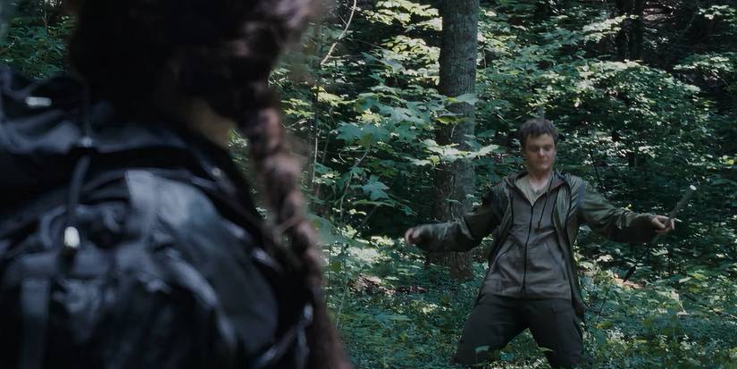 Marvel, played by Jack Quaid, falls onto his knees after being shot with an arrow by Katniss, played by Jennifer Lawrence. Marvel is wearing a green jacket, whilst Katniss wears the same jacket in black.