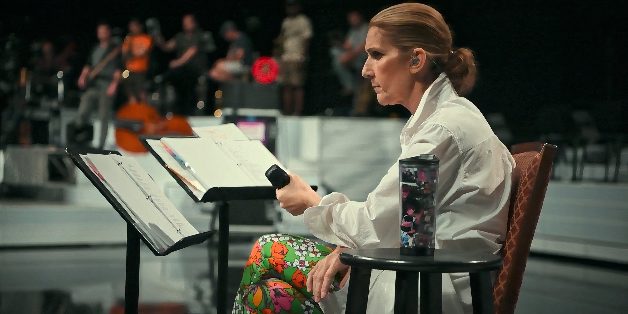 Celine Dion sits behind music stands and holds a microphone in her hand. 