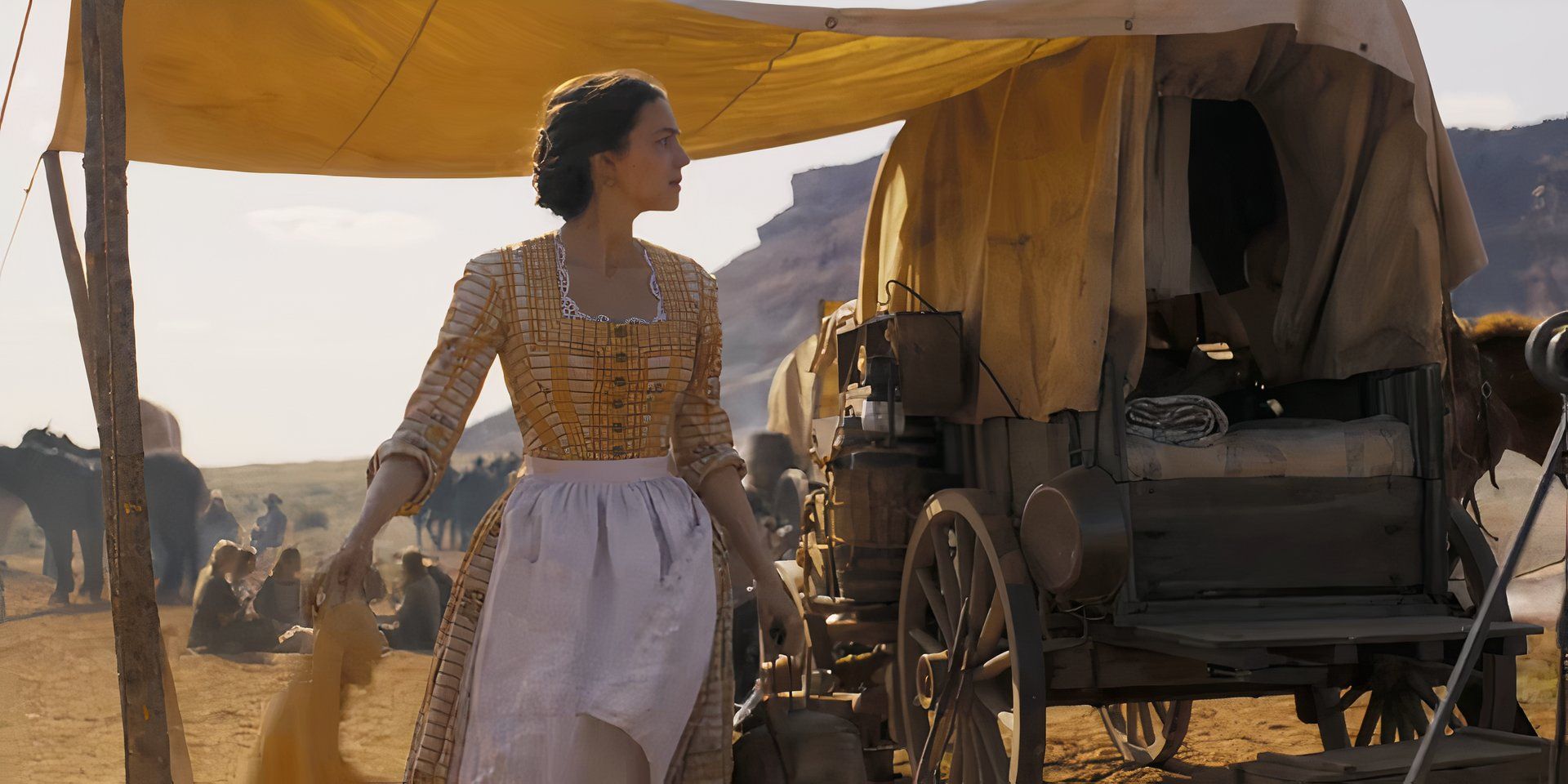 Ella Hunt as Juliette Chesney wearing an apron and holding a cloth next to a covered wagon in Horizon: An American Saga - Chapter 1