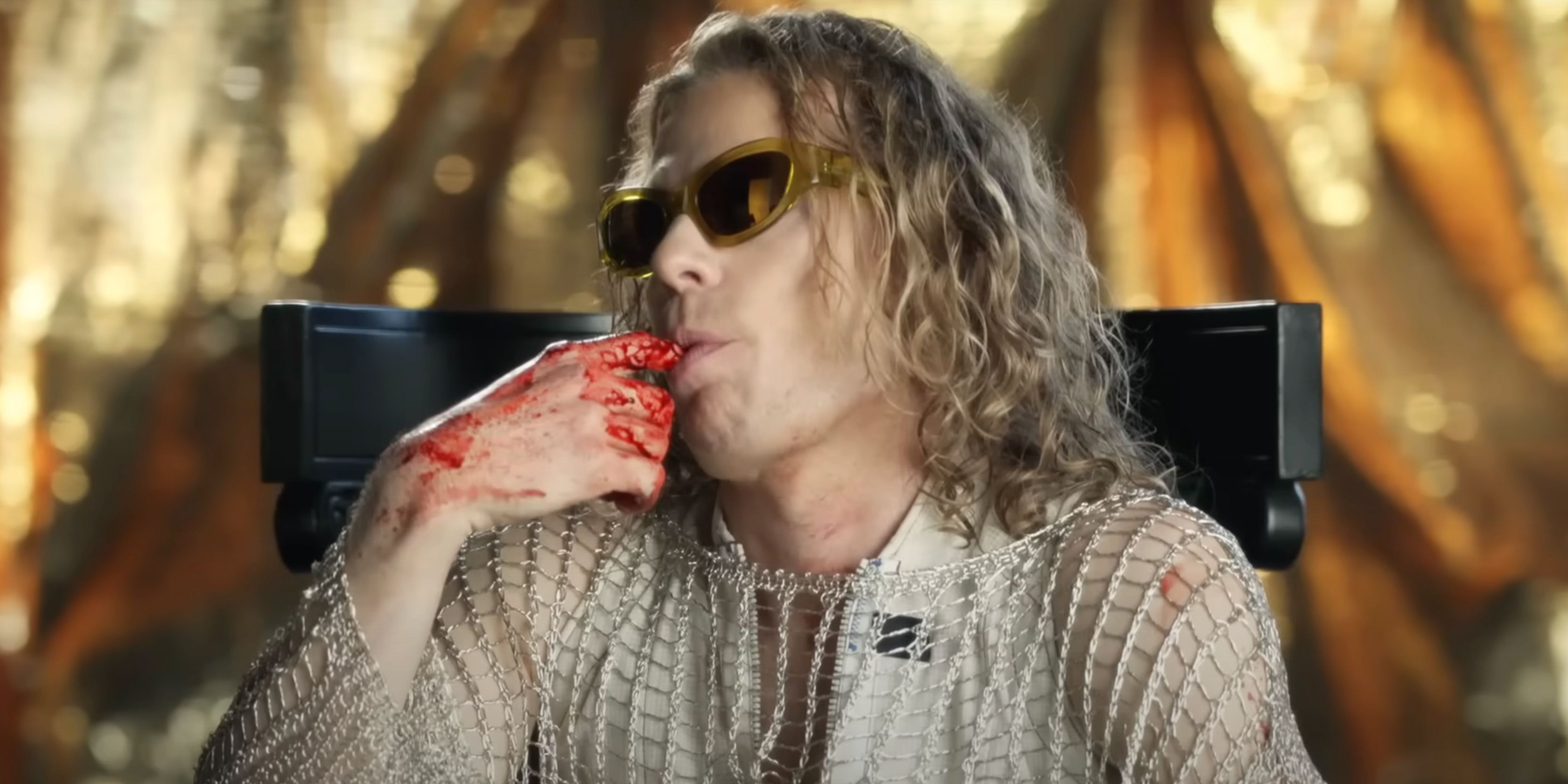 Sam Reid as Lestat licks blood off of his finger in sunglasses and a chainmail shirt in Interview with the Vampire.