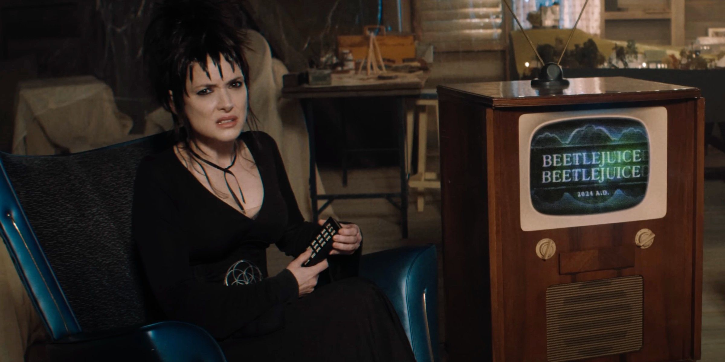 Lydia Deetz (Winona Ryder) segurando um controle remoto e sentada perto de uma TV com o logotipo do Beetlejuice 2 nela