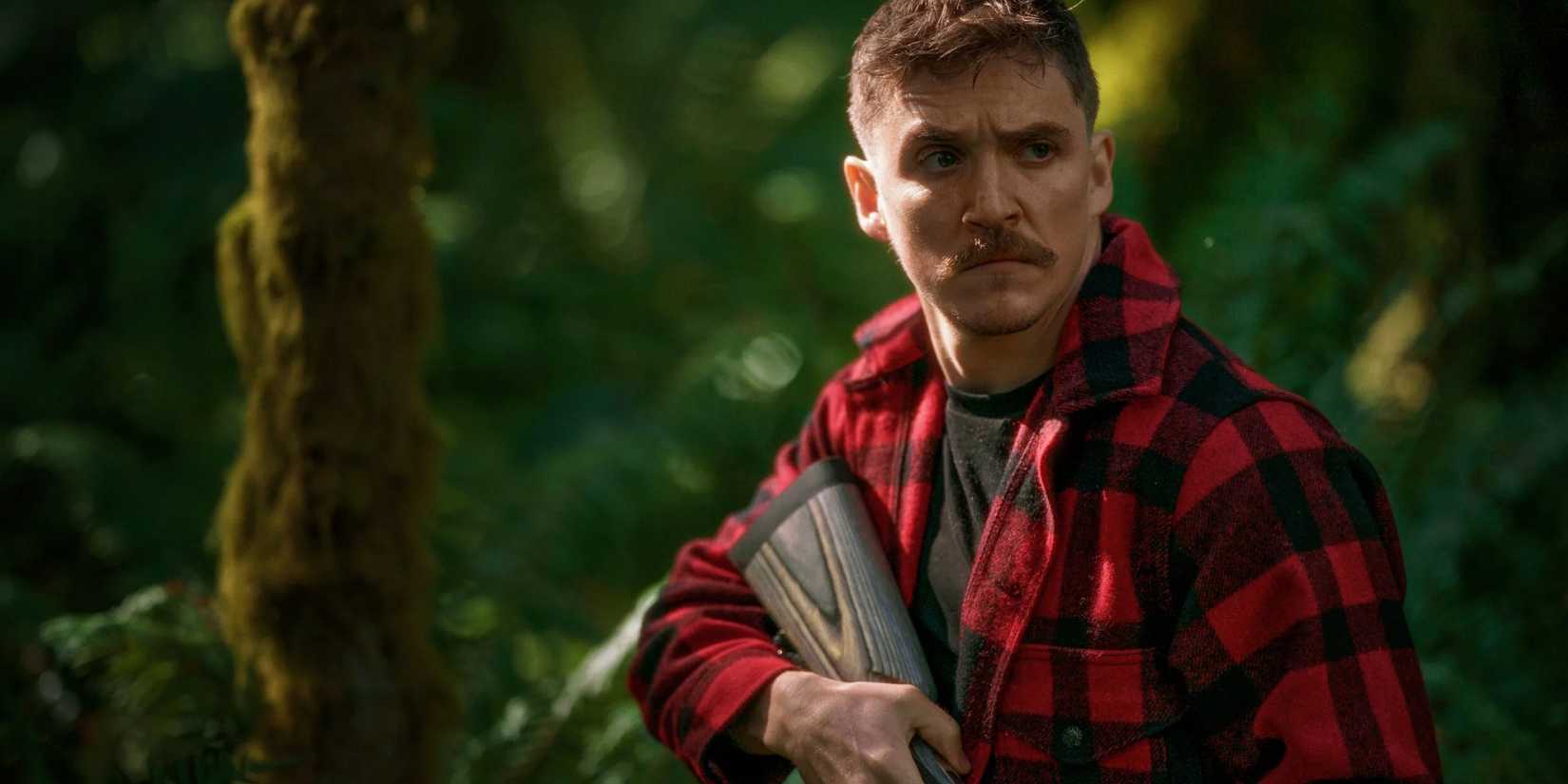 Kyle Gallner holding a rifle, wearing red and black flannel, and standing in the forest in Strange Darling