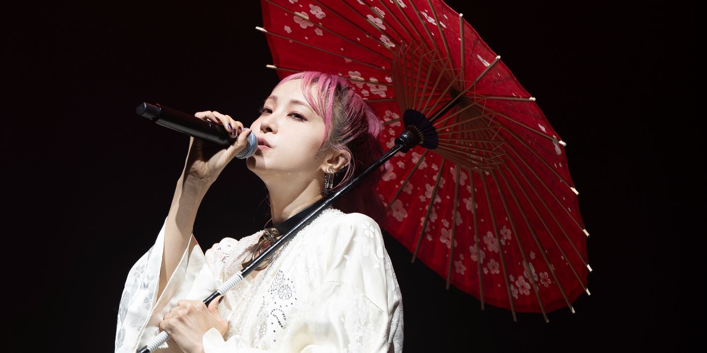 A musicista japonesa LiSA em um show vestindo um quimono com um guarda-chuva em uma mão e um microfone na outra.