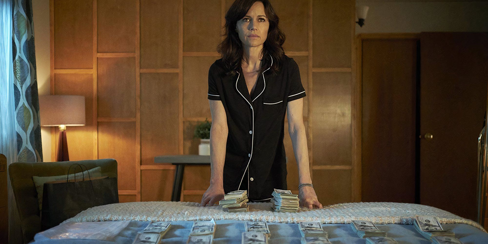 Carla Gugino as Daisy "Jett" Kowalski wearing pyjamas and stood over a bed covered in piles of money in Jett.