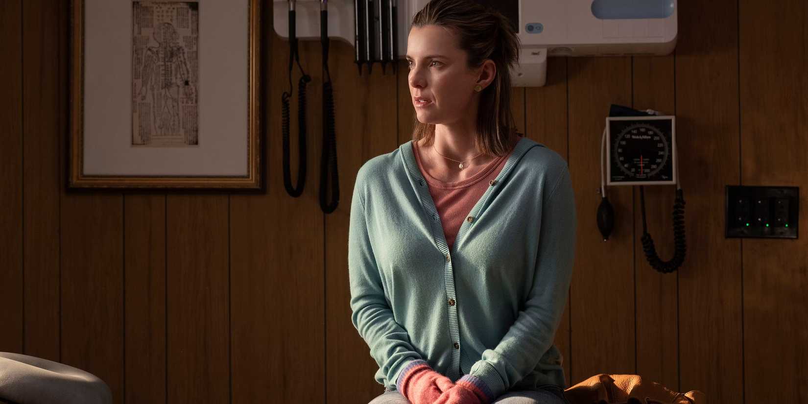 Lina (Betty Gilpin) sitting on the bed in a rustic doctor's office for Three Women