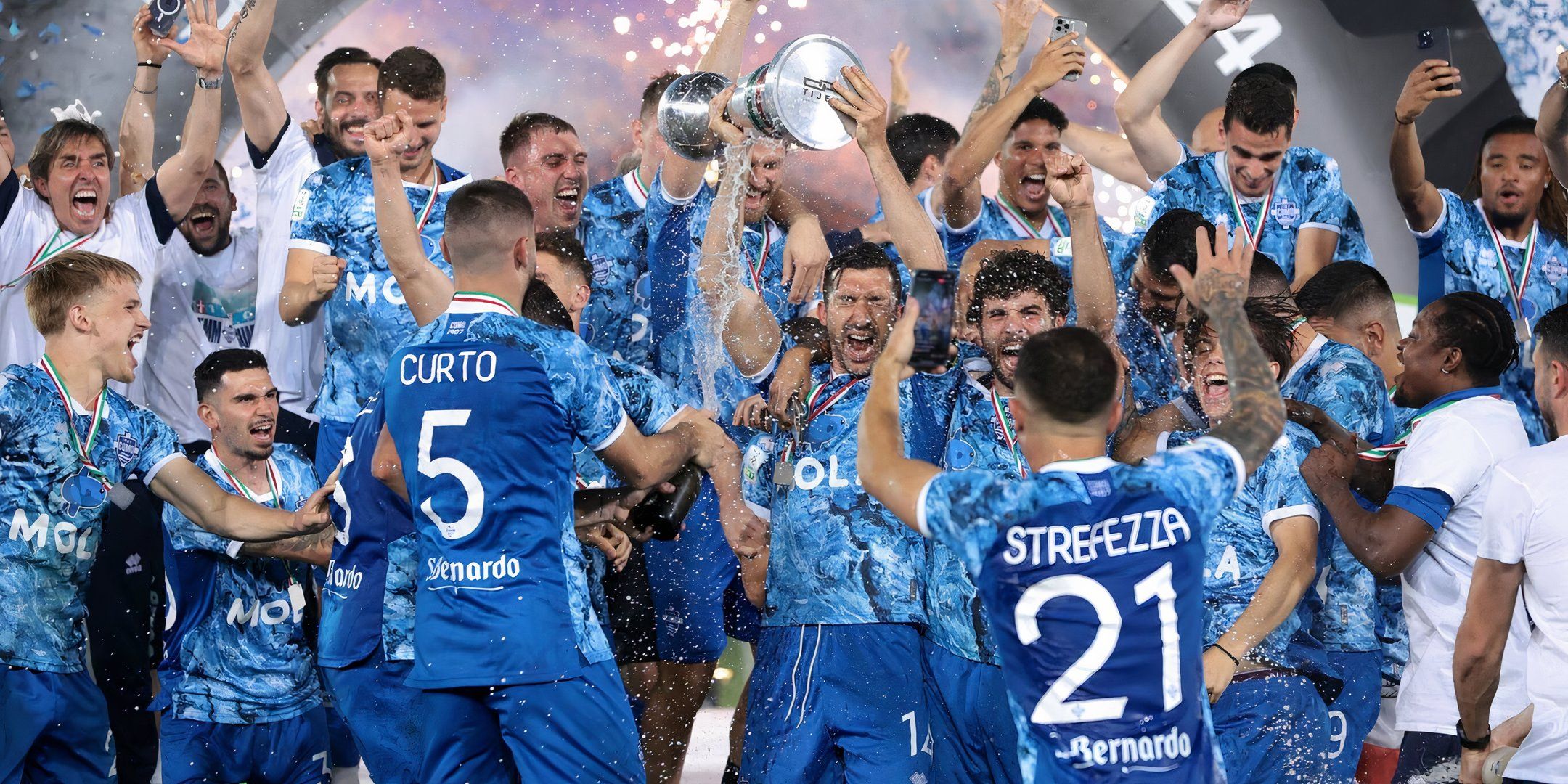 Como 1907 players celebrate the victory as they lift the trophy. The team, wearing their blue and white camouflage jerseys, is surrounded by teammates and staff.