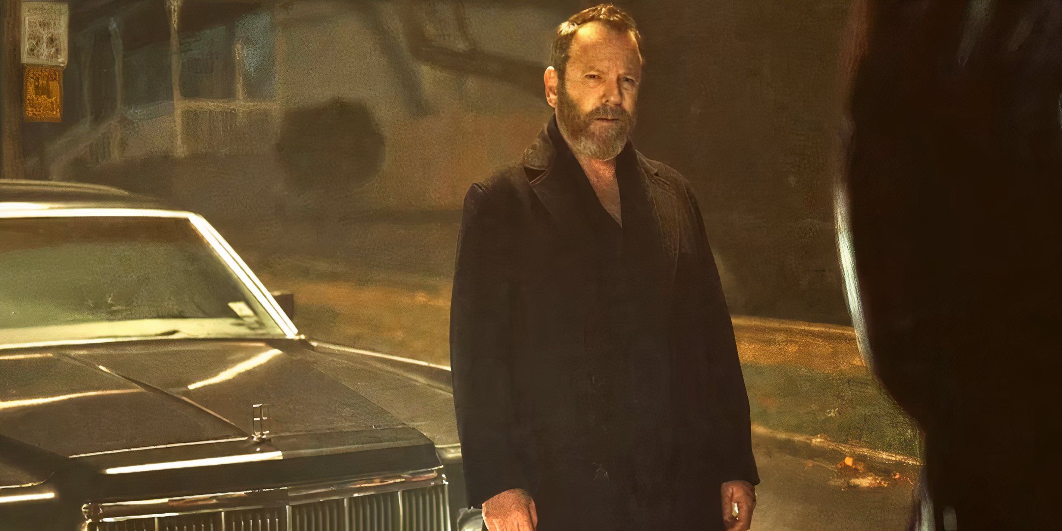 Kiefer Sutherland está parado na frente de um carro e semicerra os olhos para as pessoas à sua frente.