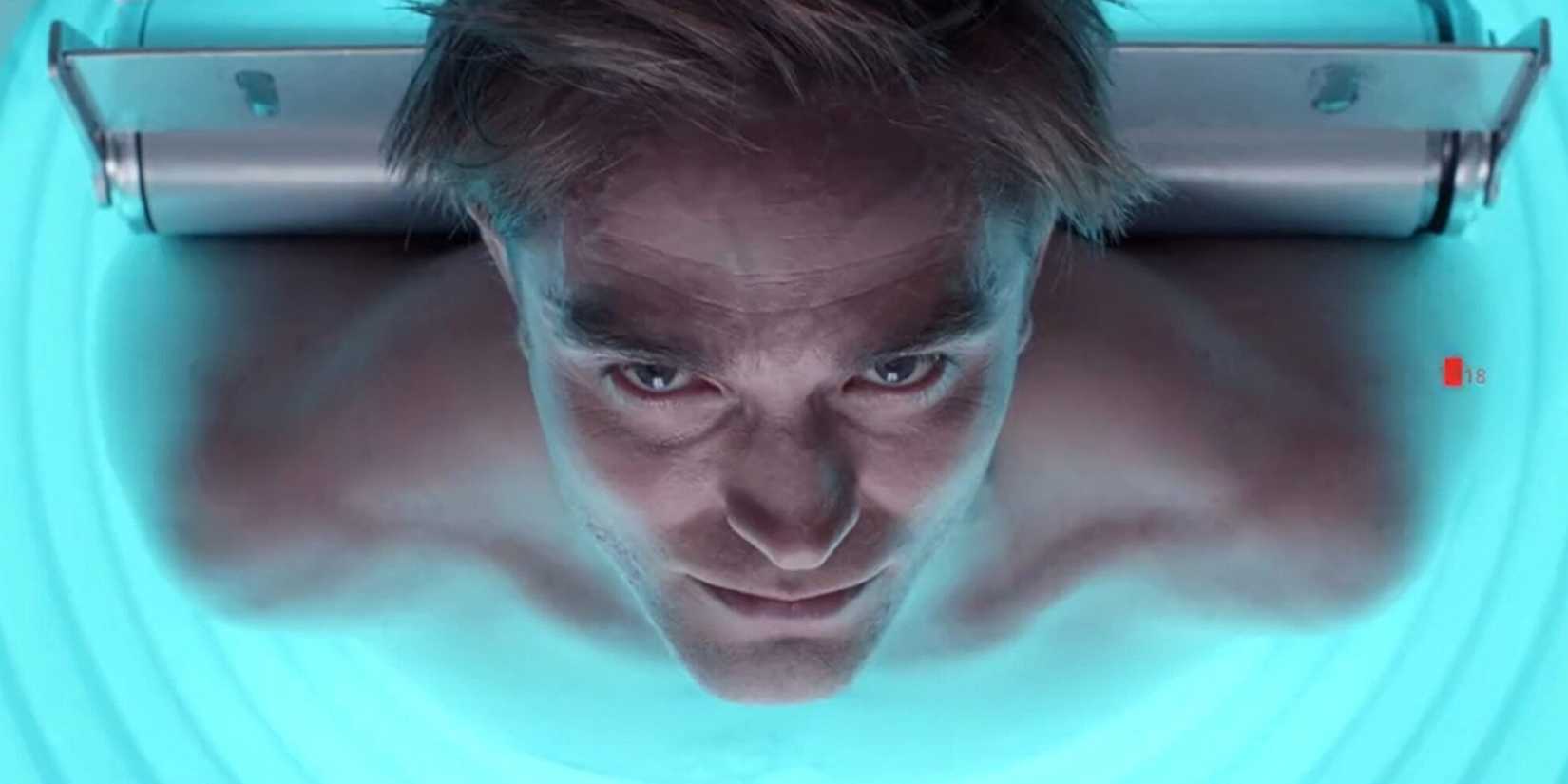 Robert Pattinson lying on a slab with his head hanging upside down and staring into the camera for Mickey 17