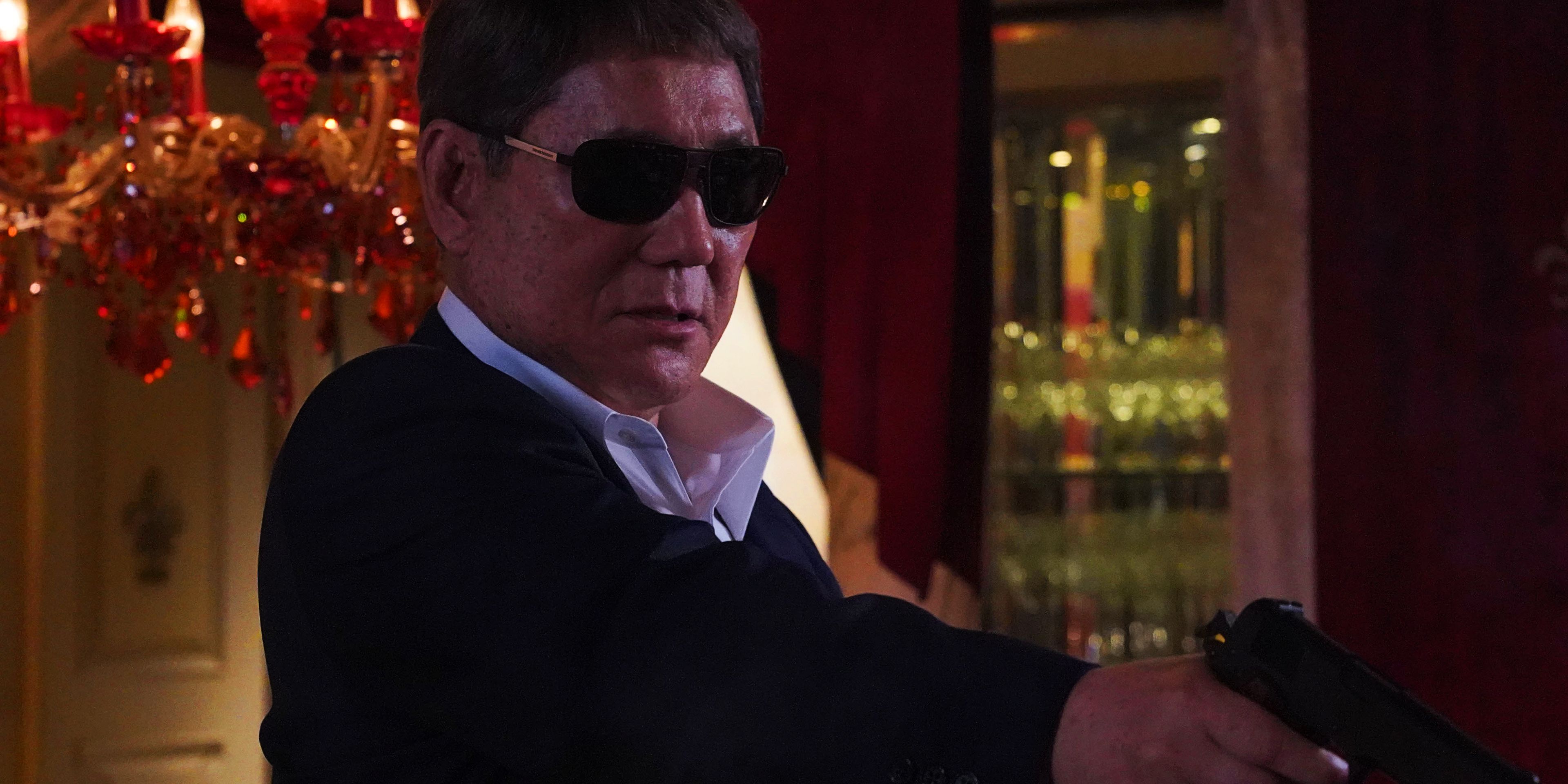 Takeshi Kitano mengenakan kacamata hitam dan mengarahkan pistol di Broken Rage